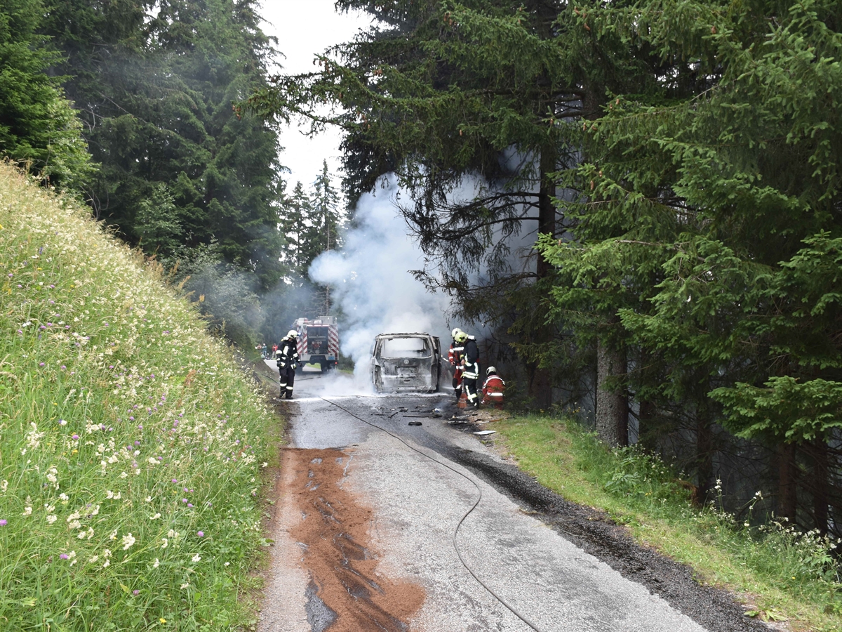 Das ausgebrannte Auto, Rauch steigt auf, rundum sind Bäume. Rund um das Auto sind Feuerwehrleute und im Hintergrund ihr Fahrzeug. 