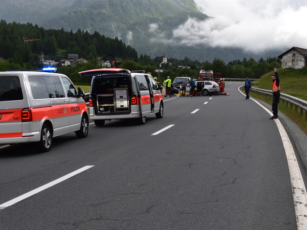 Unfallstelle auf der Malojastrasse. Am linken Bildrand zwei Patrouillenfahrzeuge der Polizei. Im Hintergrund die Unfallfahrzeuge mit Einsatzkräften der Rettung, Feuerwehr und Rega. Ein Polizist regelt den Verkehr.