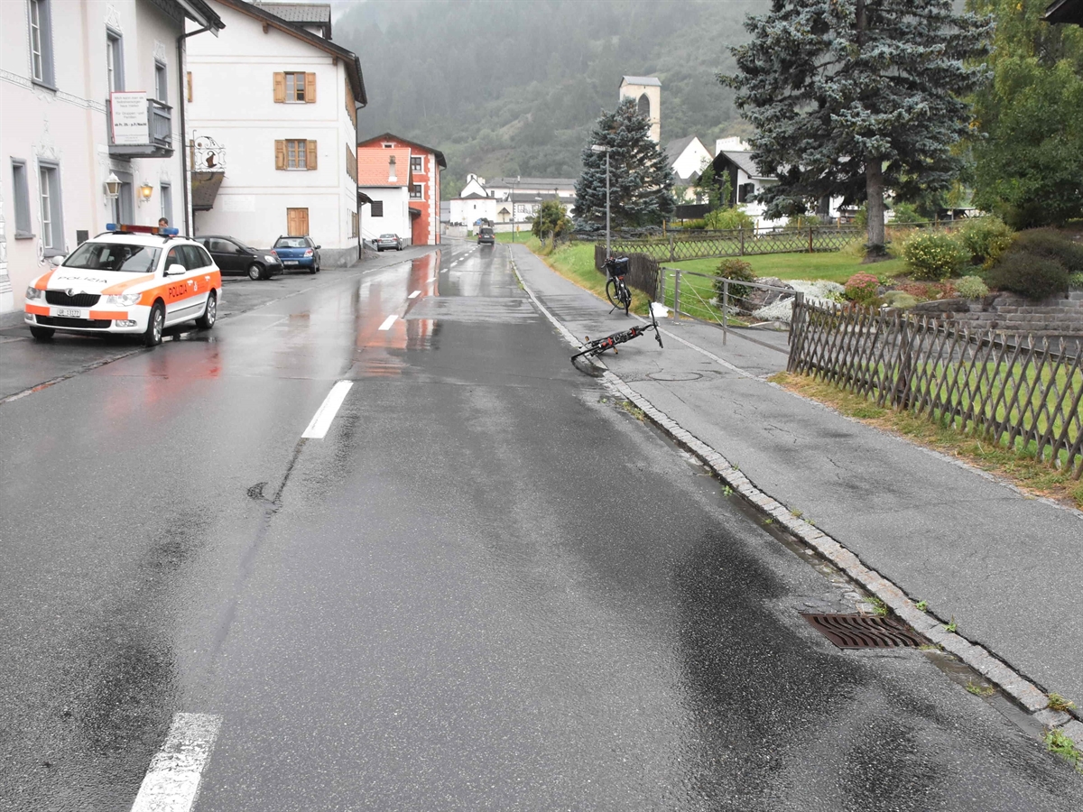 Die nasse Hauptstrasse in Richtung Dorfzentrum. Links an der Strasse ein Polizeiauto, rechts auf dem Trottoir liegt das Fahrrad.