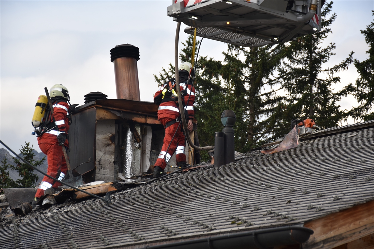 Auf dem Dach stehen zwei Feuerwehrleute im Einsatz und die Kaminabdeckung sowie Teile vom Dache wurden entfernt. Am Bildrand oben ist der untere Teil der Hubretterleiter zu sehen.