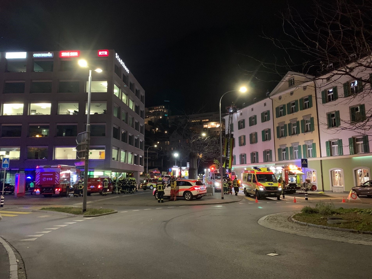 Aufnahme über einen Verkehrskreisel hinweg: Im Hintergrund rechts eine Häuserzeile, in der Mitte Einsatzfahrzeuge / Einsatzkräfte von Blaulichtorganisationen. Links davon ein weiteres Gebäude.