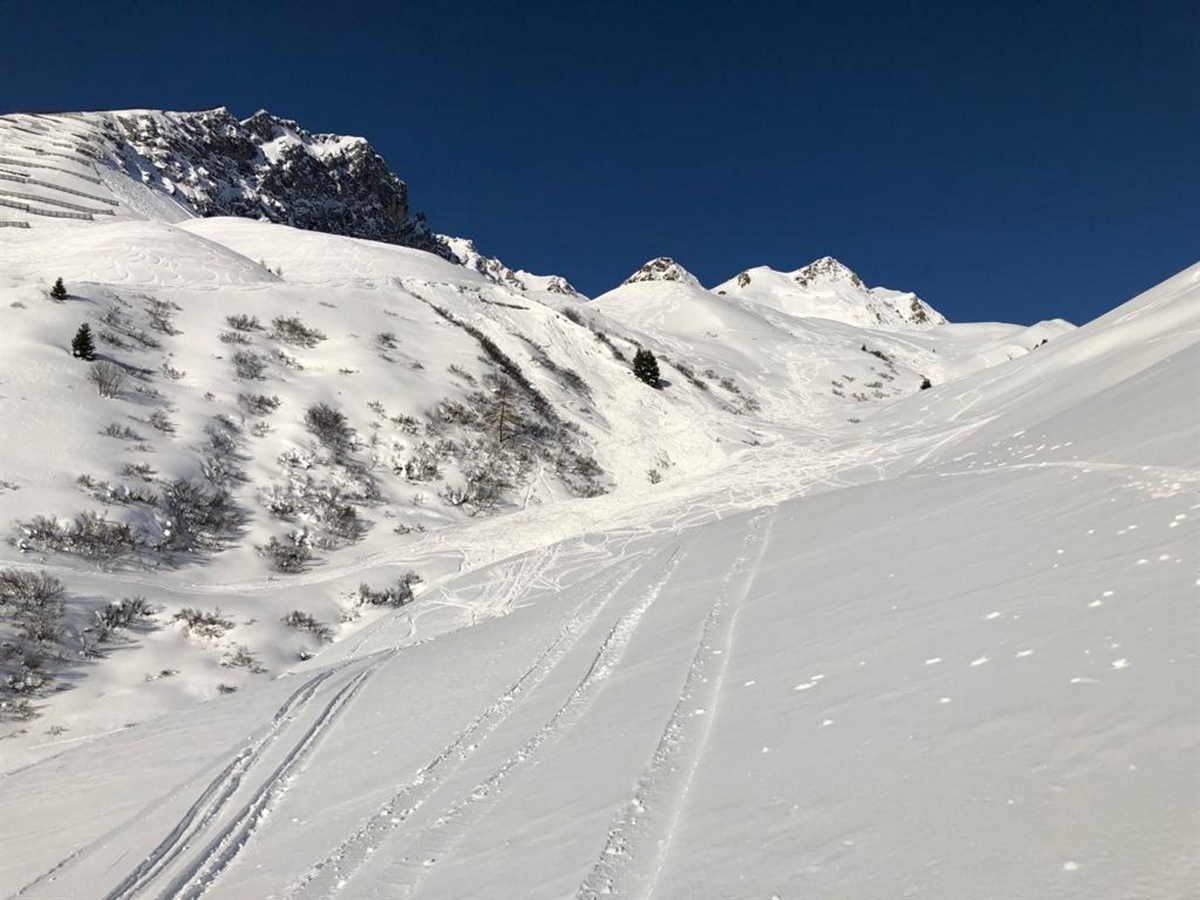 Verschneite Berglandschaft mit blauem Himmel. Mitten im Bild das Schneebrett mit vielen Skispuren im und rund um das Schneebrettfeld.