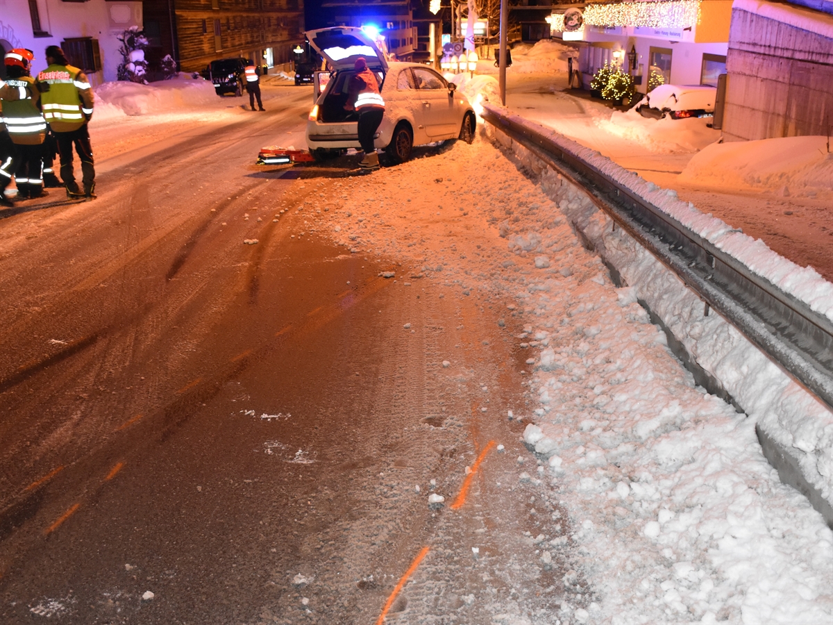 Auf der verschneiten Strasse sieht man die Schleuderspuren und rechts steht das Unfallauto an der Leitplanke. Im Hintergrund ein Feuerwehrauto und links davon ein Polizist. Weiter links Feuerwehrleute.
