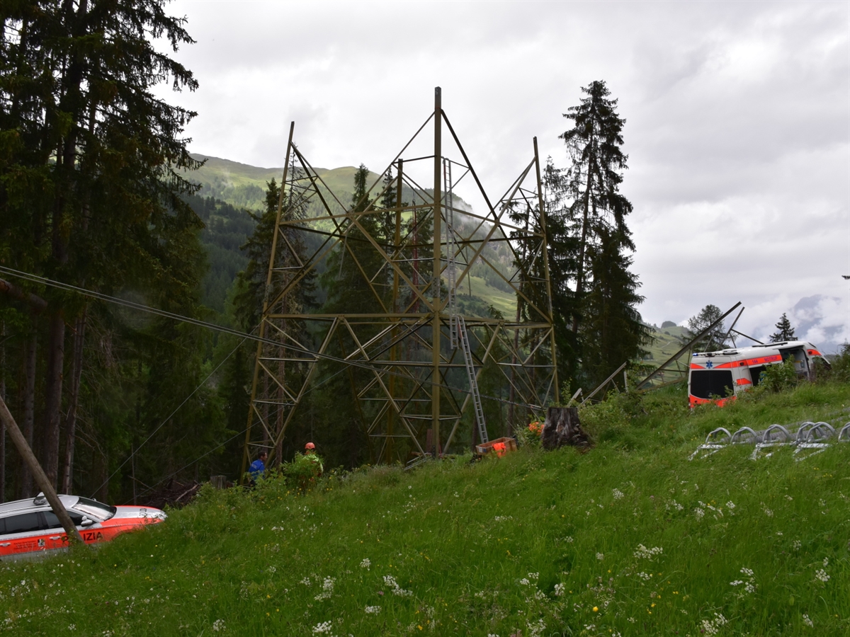 Der teildemontierte Strommast auf einer Wiese. Links ein Polizei- und rechts ein Ambulanzfahrzeug. Im Hintergrund Bäume und Berge, die den Horizont abgrenzen.