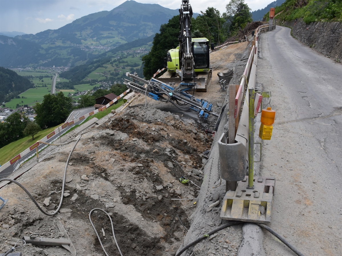 Auf der rechten Bildseite der Verlauf der Strasse in Richtung Stels. Links davon die Baustelle mit einer Baumaschine mit Bohraufsatz für Ankerbohrungen. Neben der Baustelle öffnet sich der Blick ins Vorderprättigau.