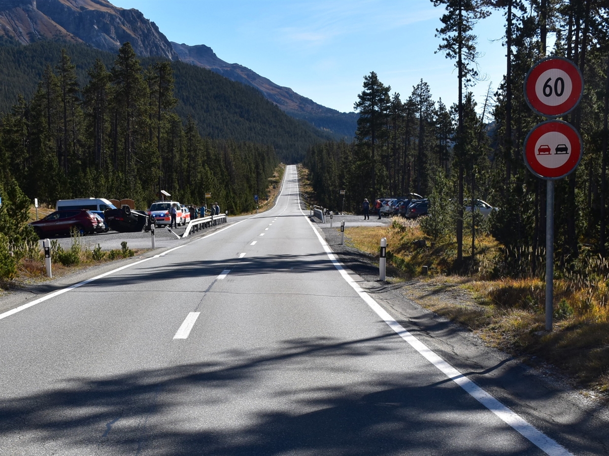 Mittig die Hauptstrasse. Rechts ein Parkplatz und die Signale 60 sowie Überholverbot. Links ebenfalls ein Parkplatz mit dem überschlagenen Auto, Personen und zwei Polizeiautos. Der Horizont wird durch blauen Himmel abgegrenzt.
