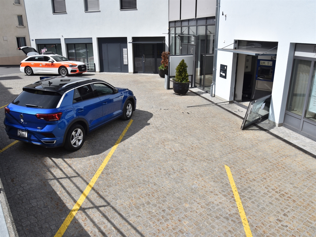 Der Vorplatz zum Bankomaten mit der beschädigten Türe zum Eingangsbereich. Auf dem Platz steht das Unfallauto sowie im Hintergrund ein Patrouillenfahrzeug der Kantonspolizei Graubünden.