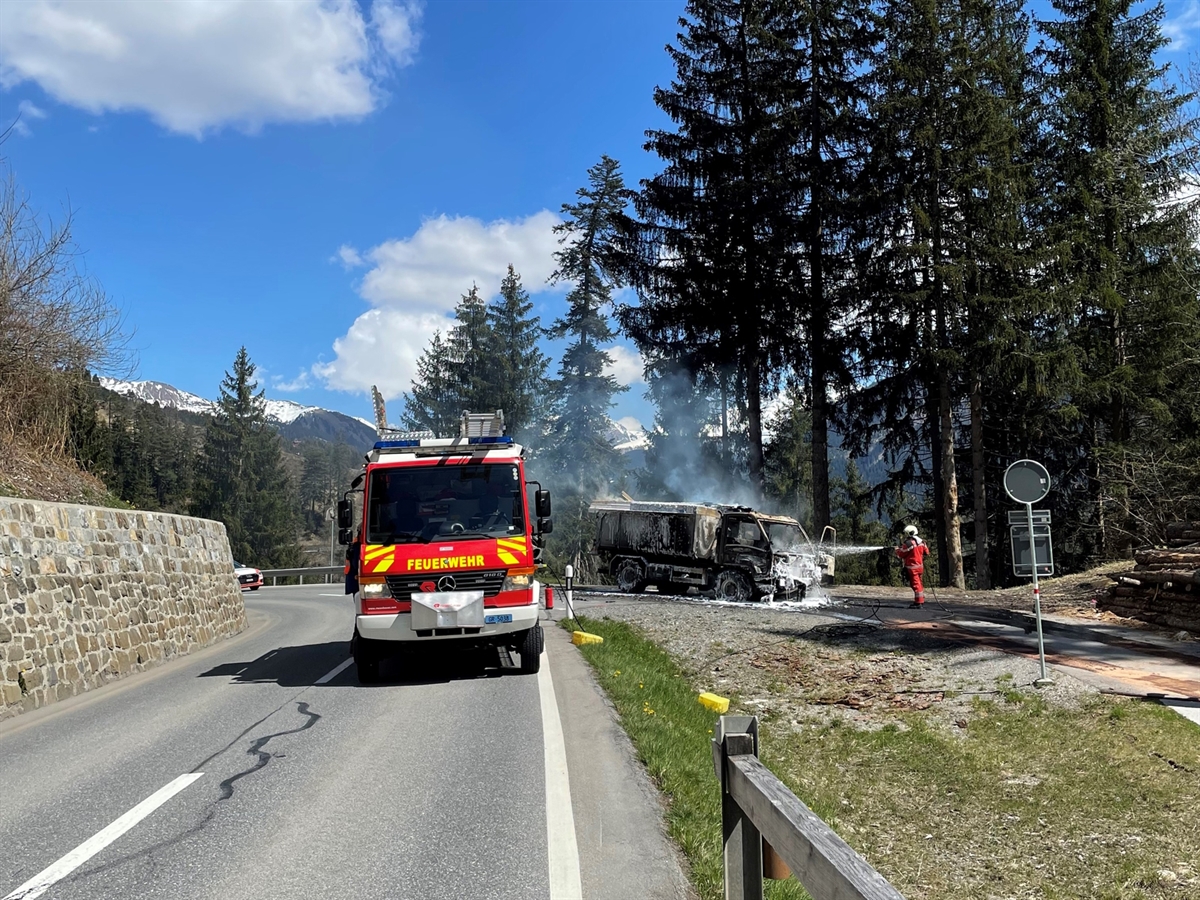 Auf der Schanfiggerstrasse steht ein Feuerwehrauto. Rechts dahinter der ausgebrannte Lastwagen. Daneben steht ein Feuerwehrmann und spritzt Löschwasser auf den Lastwagen. Links neben dem Feuerwehrauto guckt ein Patrouillenfahrzeug hinter einer Kurve hervor. Das stimmungsvolle Bild wird mit einem blauen Himmel ergänzt.