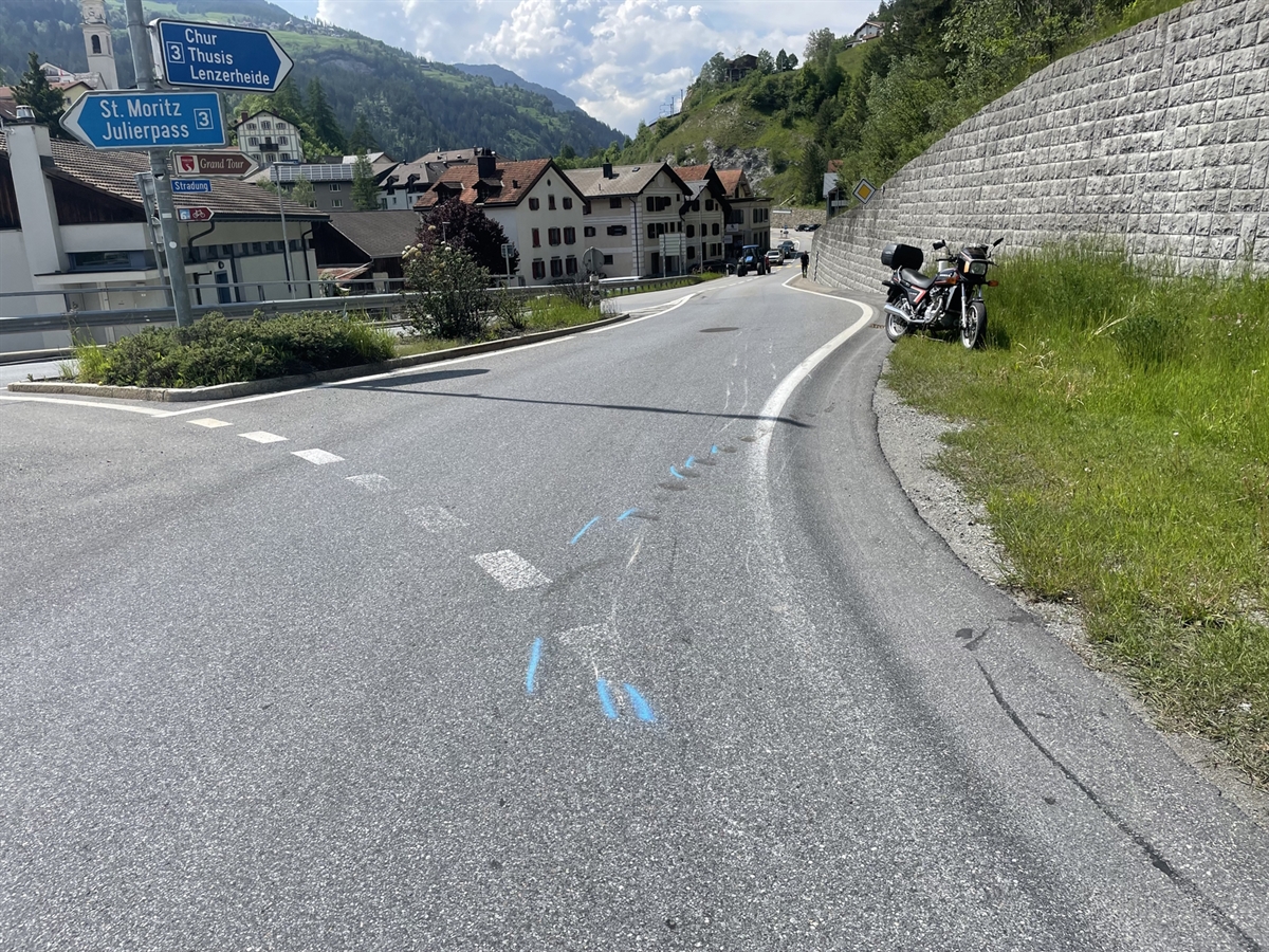 Die Ausfahrt vom Kreisel in Richtung Tiefencastel. Auf der Fahrbahn sind Schleuder und Kratzspuren des Motorrades ersichtlich. Am rechten Bildrand steht das Motorrad auf einer Wiese. Im Hintergrund Häuser von Tiefencastel