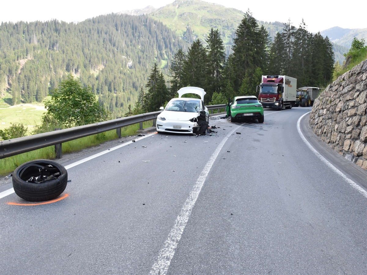 Auf der Prättigauerstrasse stehen die beiden Unfallfahrzeuge. Am unteren linken Bildrand liegt ein abgeschlagenes Rad. Hinter den Unfallfahrzeugen stehen ein Lastwagen sowie ein Traktor mit Anhänger.