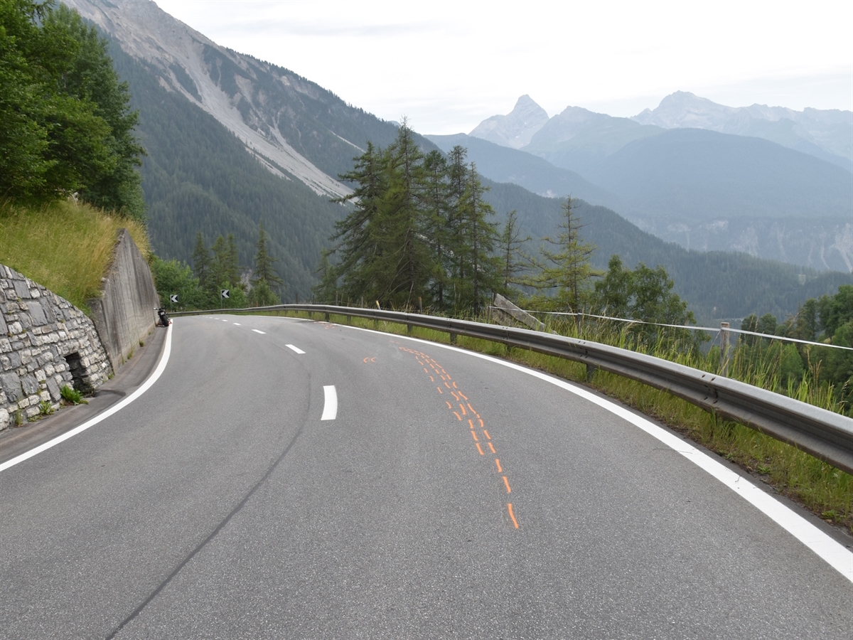 In der Linkskurve mit oranger Farbe markierte Kratzspuren des Motorrads. Dieses steht links an einer Stützmauer.