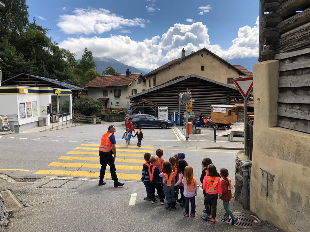 Kinder mit orangem Leuchtgürtel in einem Dorf an einem Fussgängerstreifen. Ein Polizist in Uniform weist sie an. Zwei Kinder laufen Hand in Hand über den Streifen auf die andere Strassenseite.