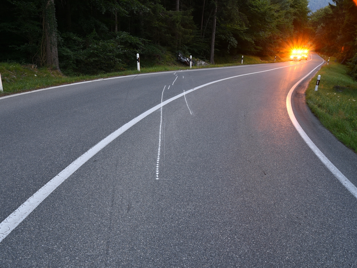 Linkskurve mit angezeichneten Kratzspuren auf der Fahrbahn. Am Strassenrand liegt das verunfallte Motorrad. Unterhalb von diesem stehen zwei Patrouillenfahrzeuge der Polizei mit eingeschalteten Warnblinklichtern