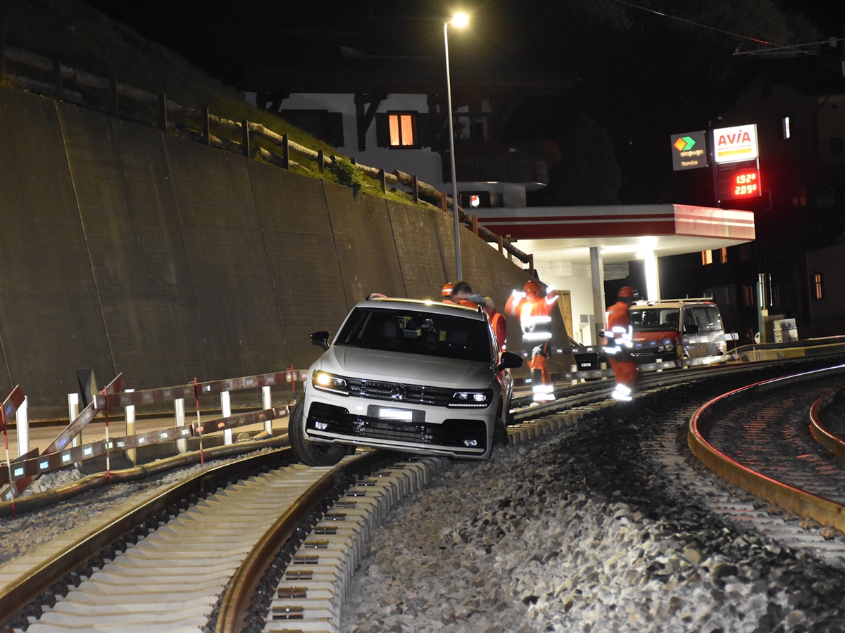 Nachtbild mit dem weissen Auto auf der Bahntrasse. Im Hintergrund Bauarbeiter sowie eine Tankstelle.