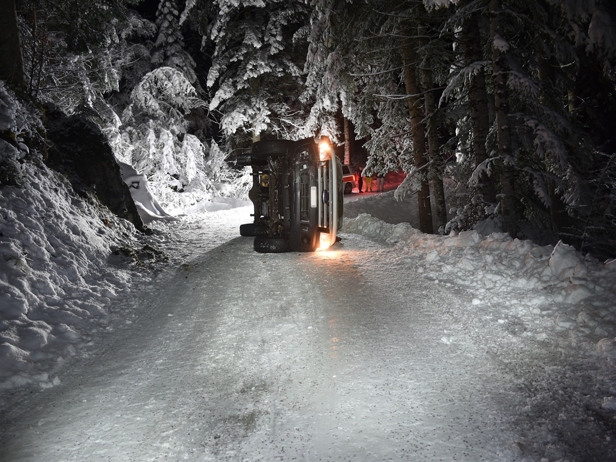 Nächtliche Winterstimmung. Verschneite Strasse, umgeben von verschneitem Wald. Auf der Strasse von vorne das auf der Fahrerseite liegende Auto. Im Hintergrund ein Polizeiauto.