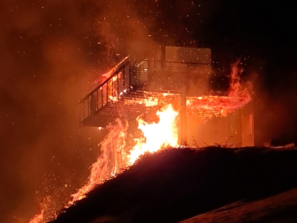 Das Bild zeigt das brennende Gebüsch und die Fassade des Ferienhauses in der dunklen Nacht