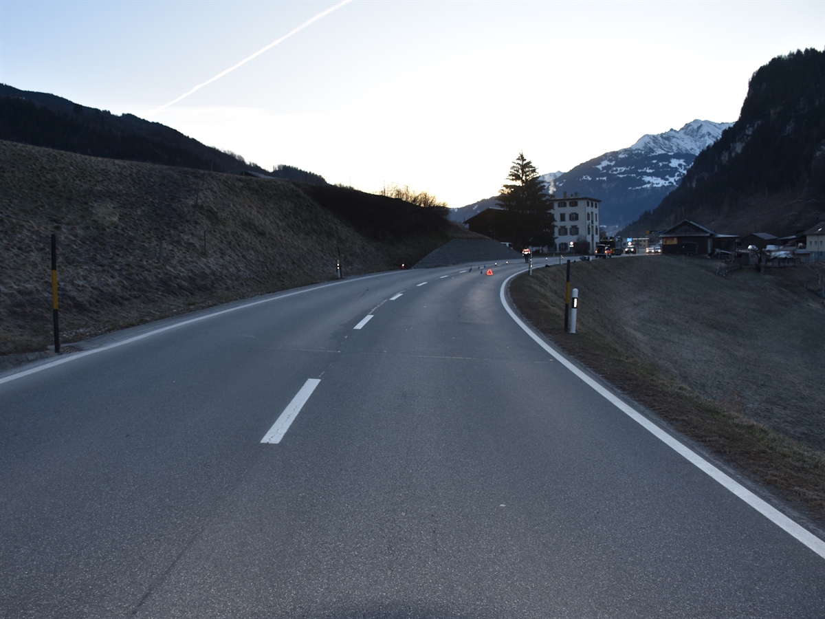 Die nach rechts verlaufende Oberalpstrasse kurz vor Dorfeingang Rueun. Auf Fahrbahn markieren Pylonen Spuren des Unfalls. Ebenfalls auf der Fahrbahn steht ein Polizist und hält den Verkehr auf. Am Horizont kündigt die aufgehende Sonne ein neuer Tag an.