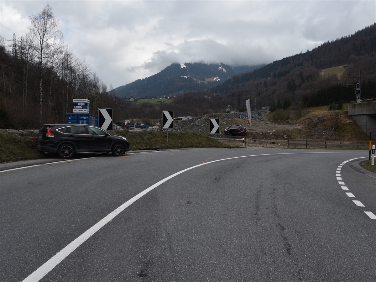 Die Rechtskurve vor der Bahnüberführung. Am linken Fahrbahnrand steht ein schwarzes Unfallauto. Im Hintergrund an einer Böschung anlehnend das andere Unfallauto.