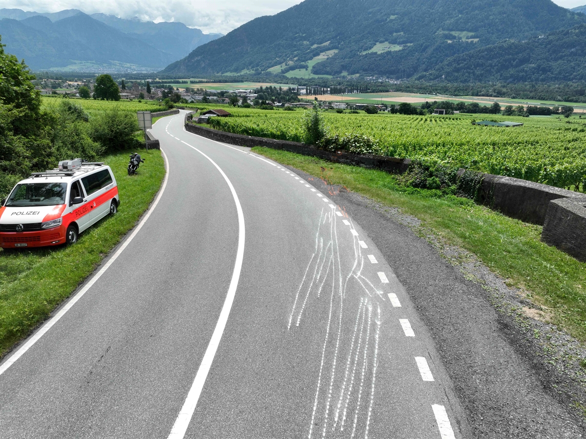 Übersicht der Unfallstelle mit Blick in Richtung Maienfeld. Links steht auf der Wiese ein Polizeifahrzeug und dahinter das Motorrad. Auf der Fahrbahn sind mit weisser Farbe die Spuren eingezeichnet. Auf der rechten Seite ist die Mauer zu sehen.