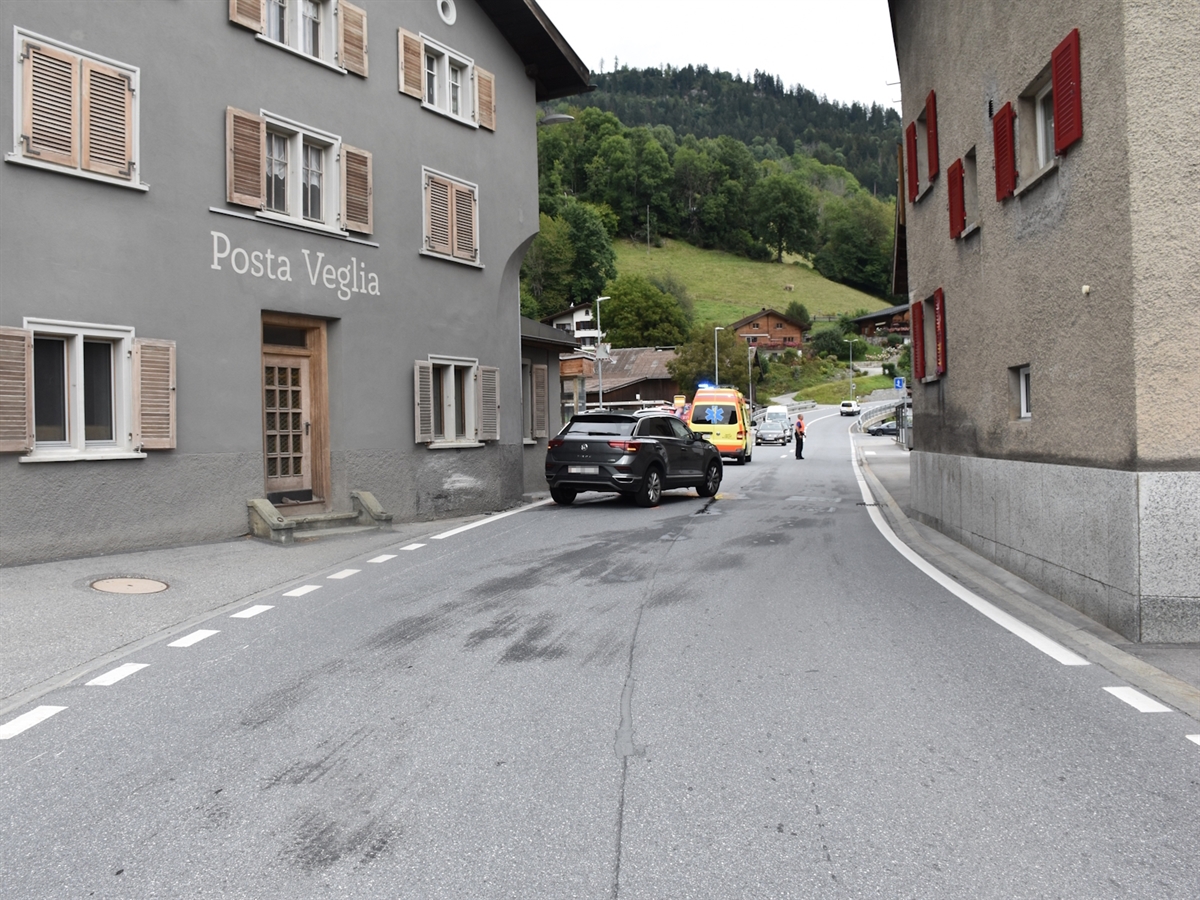 Das Auto steht im Engpass in Schluein mitten auf der Strasse. An der Hausmauer sind Kollisions- und Abriebspuren erkennbar. Hinter dem Auto stehen der Rettungswagen sowie ein Polizist auf der Fahrbahn.