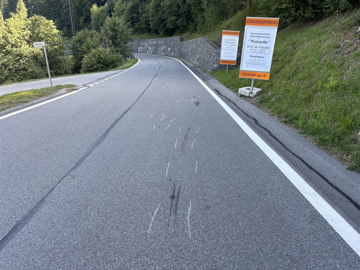Die Rechtsrheinische Oberländerstrasse in Fahrtrichtung Bonaduz gesehen. Am rechten Fahrbahnrand stehen zwei Informationstafeln über eine Strassensperrung. Auf der Fahrbahn sind Kratz- und Bremsspuren der Motorräder eingezeichnet.