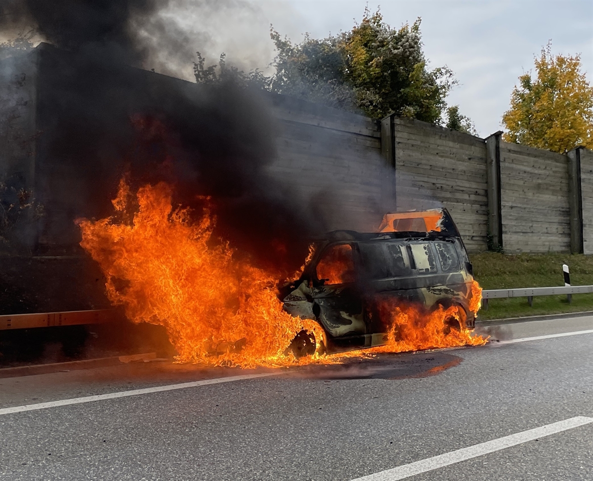 Der brennende VW-Lieferwagen auf dem Pannenstreifen der Nordspur der Autobahn A13. Hinter dem brennenden Fahrzeug die Böschung sowie die Holzlärmschutzwand. Herbstlich gefärbte Bäume ragen in den Horizont.